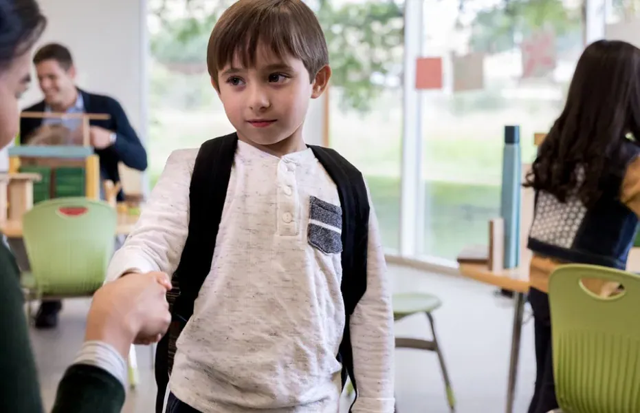 School aged child in classroom setting has uncertain facial expression, clasps parent’s hand