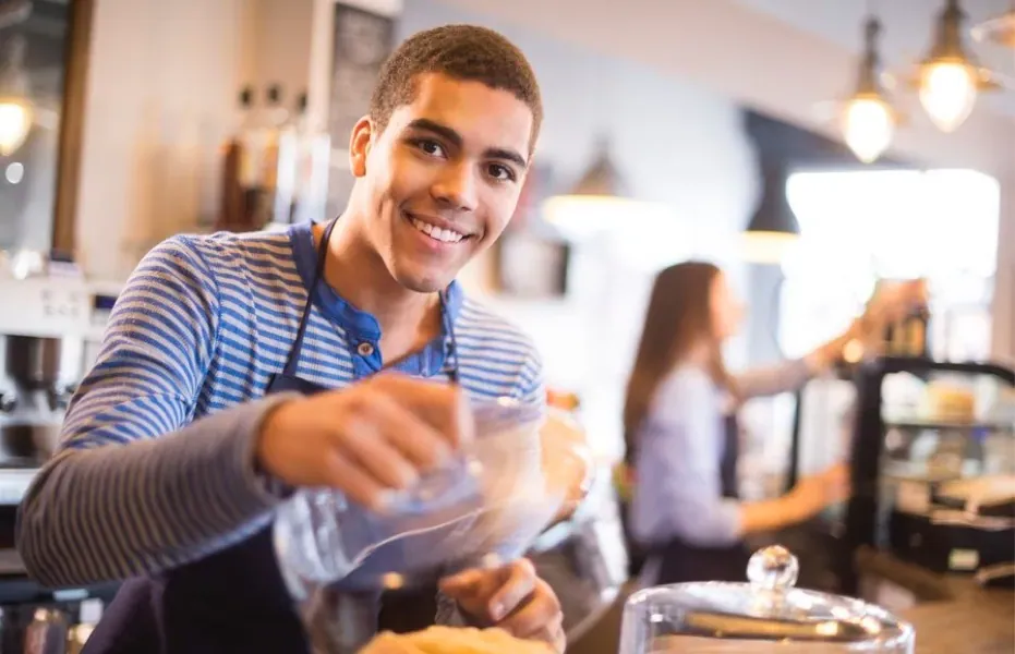 A teenager in an apron lifts a glass cover off of a tray of cookies in a restaurant.