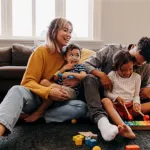 A young family sits on the floor, laughing and playing together with toys in a cosy living room.