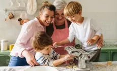 A family with two young boys making pasta together in the kitchen, guided by their mother and grandmother.