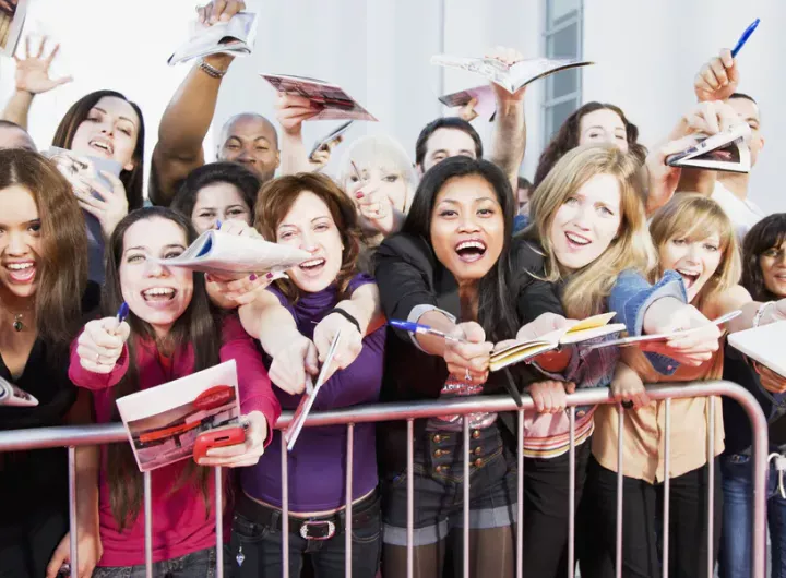 crowd of fans behind a fence screaming and waving autograph books